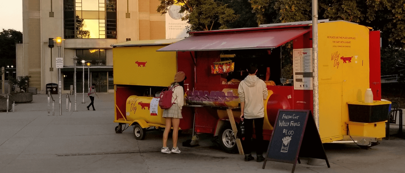 Students standing near red and yellow campus hot dog stand on wheels with awning